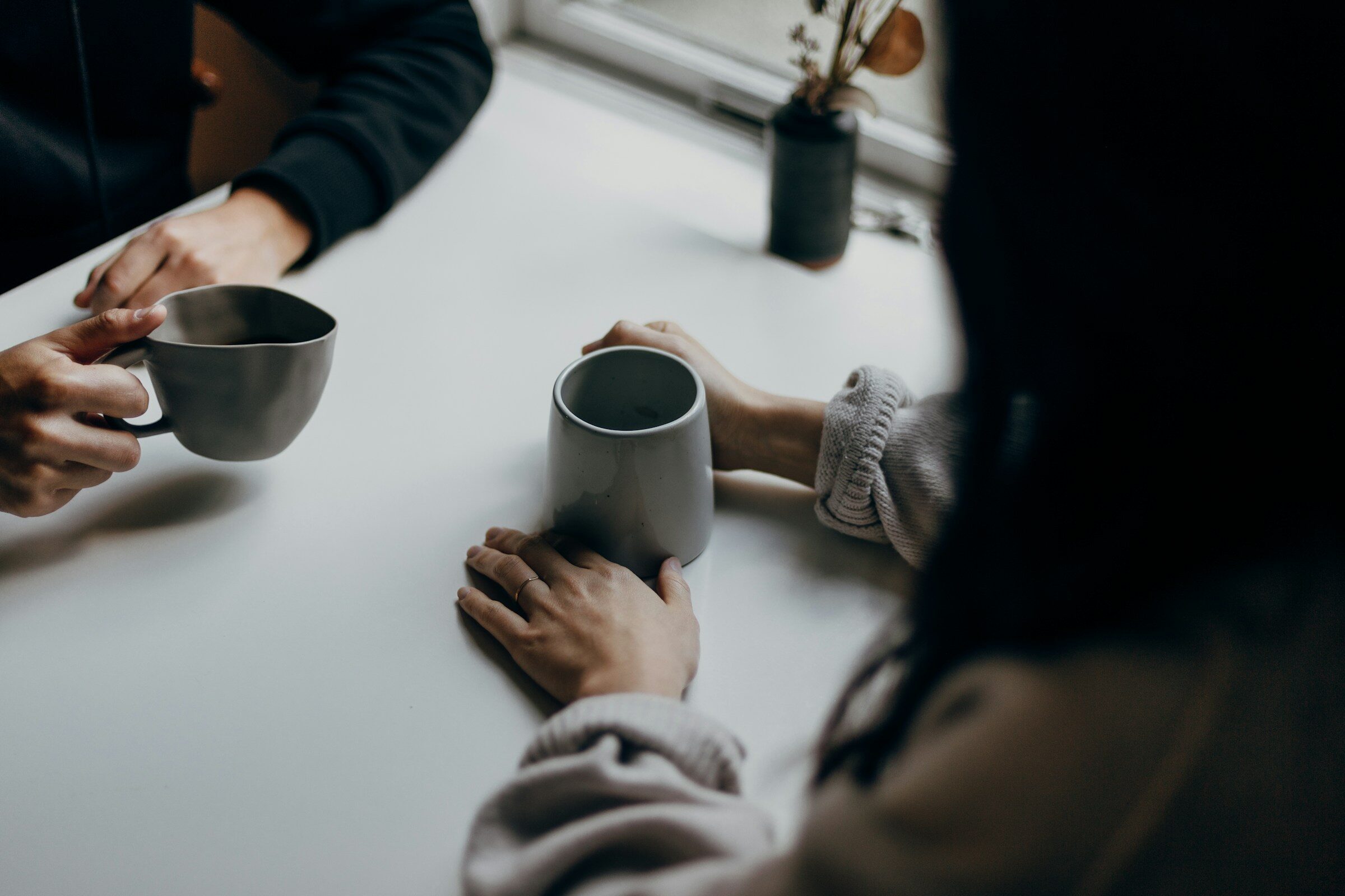 Two people talking over coffee, hands round their mugs on a table