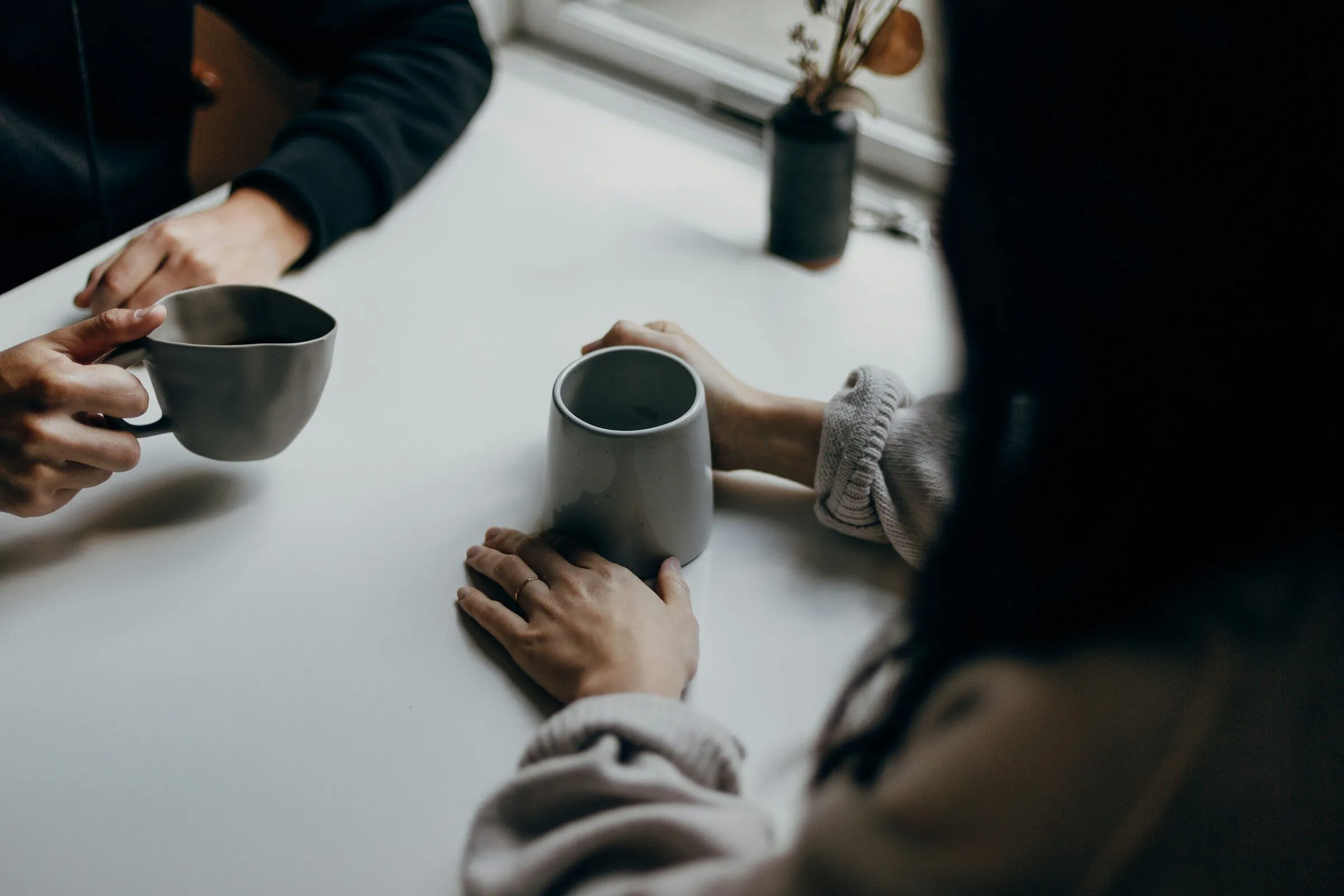 Two people talking over coffee, hands round their mugs on a table